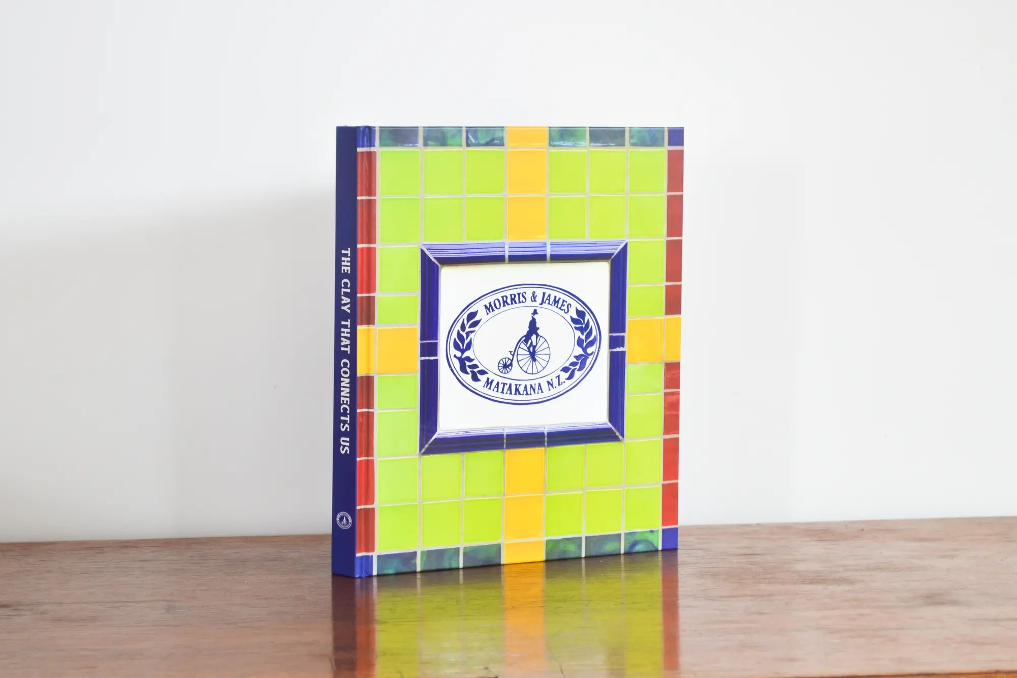 The Clay That Connects Us — the finished hardcover standing on a wooden table, showing the mosaic tile cover and the Morris & James penny-farthing mark